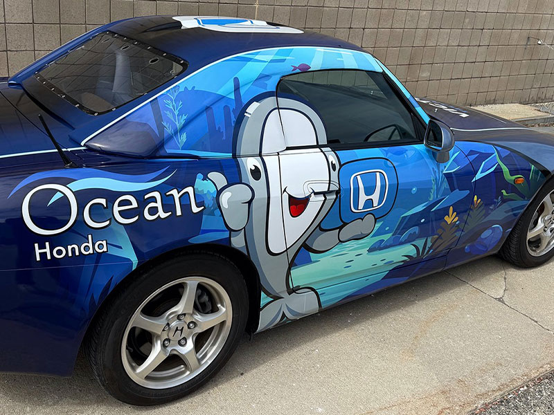 Blue Honda car with an Ocean Honda logo and cartoon fish graphic on the side, parked by a brick wall.